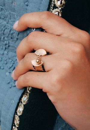 Bague de couleur or présentant un design en forme d'éventail avec des crêtes et un petit accent en forme de boule. Une main tient un tissu texturé et sombre.