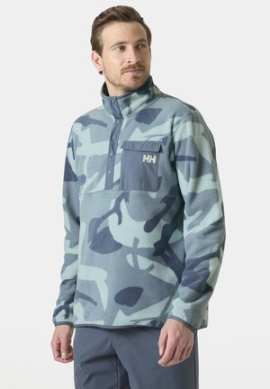 Hombre con un forro polar de patrón de camuflaje azul y gris, con cuello alto y bolsillo en el pecho, de pie y mirando hacia la derecha.