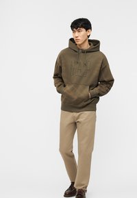 Νέος άνδρας που φοράει πράσινο-ελαιίχρωμο φούτερ GAP, μπεζ παντελόνι και καφέ παπούτσια, στέκεται με τα χέρια στις τσέπες του φούτερ, κοιτώντας προς τα πλάγια.