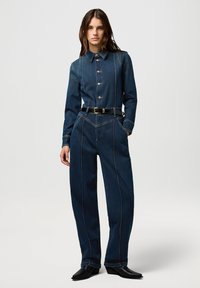 Mono de mezclilla azul oscuro con cuello, cierre de botones en la parte delantera y pantalones de pierna ancha. Presenta costuras en contraste y un cinturón negro en la cintura.