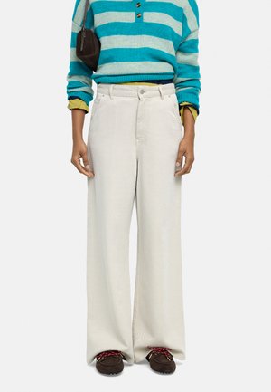 Osoba nosząca szerokie, kremowe spodnie, sweter w paski w kolorach morskim i jasnoniebieskim na żółtej koszuli, brązowe buty z czerwonymi sznurówkami oraz małą torebkę.