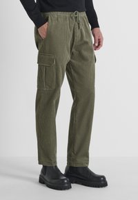 Pantalones cargo en pana verde oliva, con una cinturilla elástica y cordón, bolsillos laterales y textura acanalada. Botas negras gruesas.