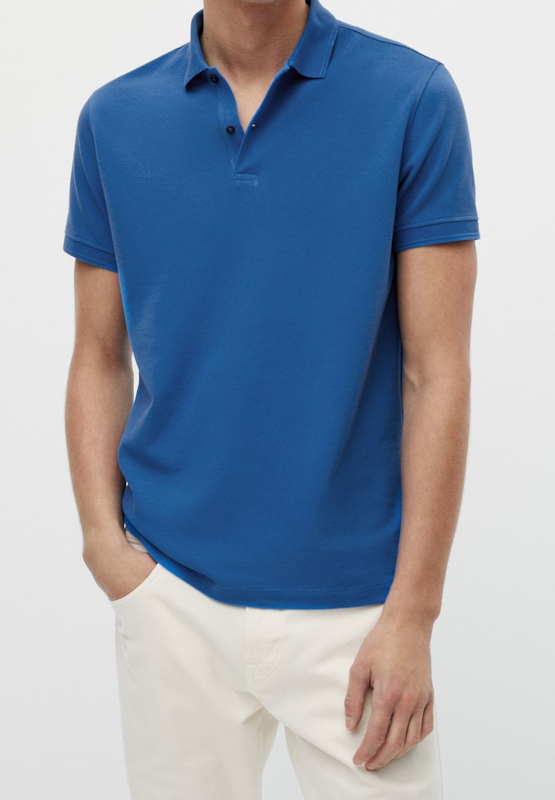 Homme portant un polo bleu à manches courtes et un pantalon blanc, debout avec une main dans la poche devant un fond uni.