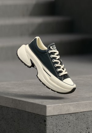 Μαύρο και άσπρο χαμηλό sneaker με παχιά σόλα, δεμένα κορδόνια, τοποθετημένο πάνω σε μια τσιμεντένια άκρη με φόντο θολό γκρι.