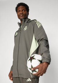Grå letvægtsjakke med mintgrønne detaljer og striber. Har en lynlås, hætte, og et Real Madrid-logo. Holder en mønstret fodbold.