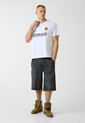 Joven de pie, viste una camiseta blanca con la palabra "ALEMANIA" y un emblema de la bandera, pantalones cortos negros hasta la rodilla, calcetines grises y botas marrones con cordones.