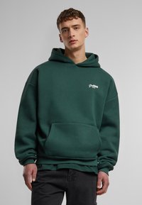 Sudadera de color verde oscuro hecha de una mezcla de algodón, con un bolsillo frontal, puños acanalados y un pequeño logo blanco en el pecho.
