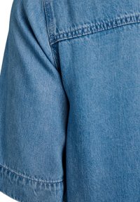 Nahaufnahme eines hellblauen Jeanshemds mit kurzen Ärmeln, das die Stoffstruktur und die Nähte an Schulter und Ärmel zeigt.