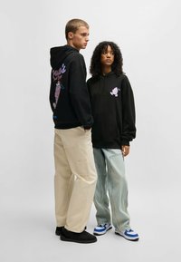 Felpe nere con stampe grafiche, vestibilità rilassata; il modello indossa pantaloni cargo color crema e jeans oversize azzurri, entrambi con cuciture visibili.