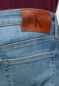 Vybledé modré rifle z denimu s hnedou koženou nášivkou s ražbou "Calvin Klein" a logom "CK", s kontrastným prešívaním a zadnými vreckami.