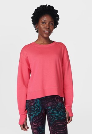 Vrouw draagt een koraalroze sweatshrit met lange mouwen en zwarte leggings met kleurrijke golvende lijnpatronen, staand tegen een effen witte achtergrond.