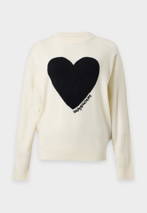Femme portant un pull blanc avec un grand cœur noir, un jean bleu, une ceinture marron, des chaussures marron et tenant un sac à main marron avec deux poignées.
