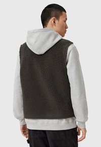 Gilet en polaire marron foncé superposé sur un sweat à capuche gris clair, présentant une surface texturée et une coupe décontractée, sans fermetures visibles.