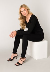 Zwarte lange mouwen top en aansluitende broek, gecombineerd met zwarte sandalen met hoge hakken en bandjes. Model zittend op een witte kubus tegen een lichte achtergrond.