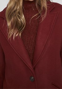 Manteau en mélange de laine bordeaux avec des revers crantés, une fermeture à un bouton et des poches latérales, superposé sur un pull à col roulé côtelé assorti.