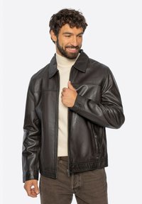 Man die glimlacht en naar beneden kijkt, gekleed in een donkerbruine leren jas over een beige coltrui en bruine broek.