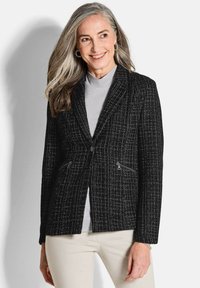 Blazer noir en tweed avec une finition texturée, deux poches zippées et une boutonnière unique. Associé à un haut gris clair et un pantalon crème.