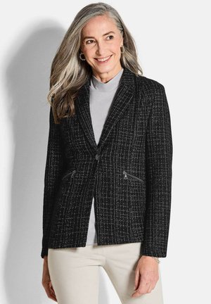 Blazer noir en tweed avec une finition texturée, deux poches zippées et une boutonnière unique. Associé à un haut gris clair et un pantalon crème.