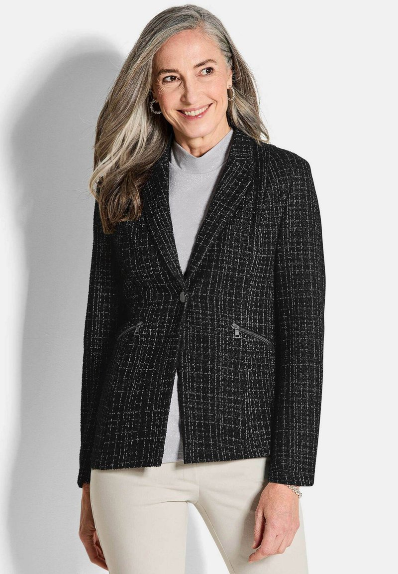 Blazer noir en tweed avec une finition texturée, deux poches zippées et une boutonnière unique. Associé à un haut gris clair et un pantalon crème.