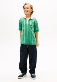 Πράσινο ριγέ πόλο πουκάμισο με λευκό γιακά, σε συνδυασμό με φαρδιά μπλε τζιν και μπλε αθλητικά παπούτσια. Εμφανές κίτρινο λογότυπο στο πουκάμισο.