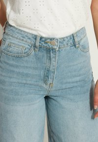 Vaqueros de tiro alto en color azul claro con cinco bolsillos, cierre de botón metálico y detalles de costura amarilla. Tela de mezclilla suave con una textura lisa.