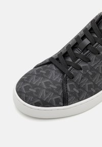 Baskets noires avec tissu texturé montrant des motifs géométriques répétitifs, lacets noirs et semelle en caoutchouc blanche.
