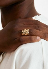 Kuldne sõrmus, millel on sile laineline disain ja läikiv viimistlus, kantud tumeda nahaga käel, mis puhkab valgel kangal.