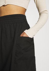 Pantalones de pierna ancha negros con cintura elástica y bolsillo lateral, combinados con una blusa ajustada de manga larga y cropped en color beige. Tela texturizada visible.