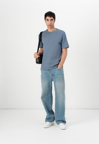 Blå bomulls t-skjorte, korte ermer, med løse lyseblå denimjeans og hvite joggesko. Modellen holder en svart skulderveske.