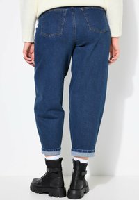 Jeans de mezclilla azul oscuro con un ajuste relajado, que presentan dobladillos enrollados y dos bolsillos traseros. Llevados con botas negras hasta el tobillo y calcetines blancos.