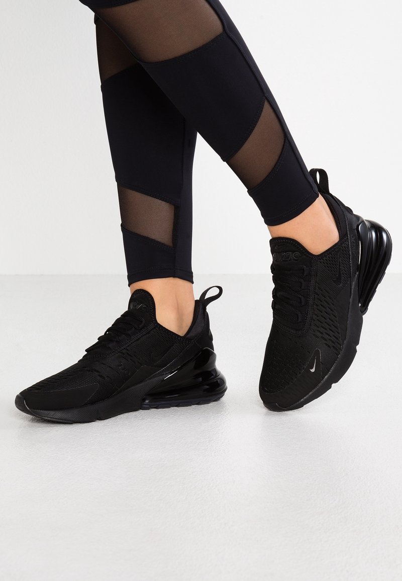 Jambes portant des leggings noirs avec panneaux en mesh et des baskets Nike Air Max 270 noires sur un sol blanc, un pied légèrement levé.