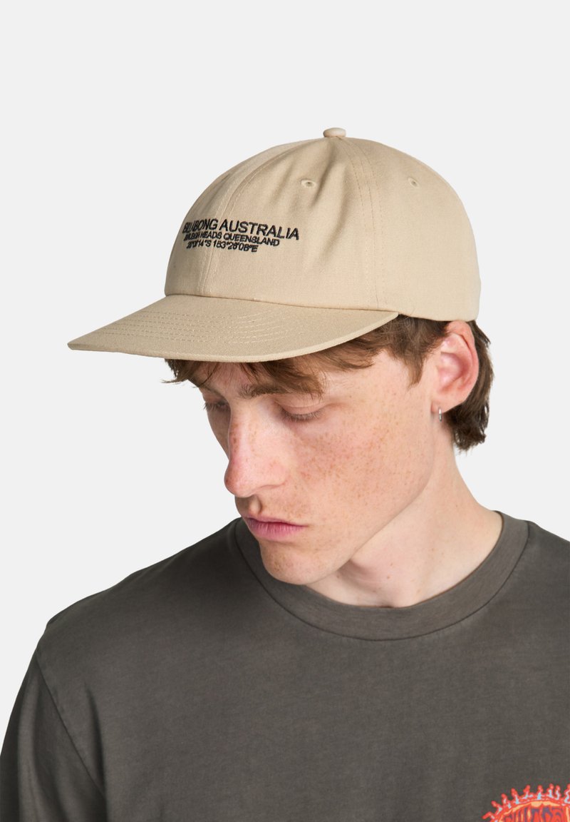 Casquette de baseball beige avec logo noir brodé, visière incurvée et design structuré. Portée légèrement inclinée, avec une texture subtile.