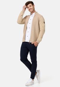 Cárdigan de punto beige con cremallera en el frente, diseño texturizado, puños acanalados, sobre una camisa blanca de botones, combinado con pantalones azul marino y zapatillas blancas.