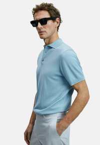 Helles blaues Poloshirt mit einem klassischen Kragen, zwei Knöpfen, kurzen Ärmeln und einer glatten Textur. Getragen mit hellen Hosen und schwarzen Sonnenbrillen.