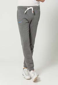 Pantalones deportivos grises con cintura ajustable, dos bolsillos laterales y puños acanalados. Incluye un pequeño logo azul y rojo en el muslo izquierdo.