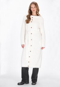 Lange witte gebreide cardigan met een riem, voorzien van grote bruine knopen en ribgebreide accenten op de hals en mouwen. Gecombineerd met donkere jeans.