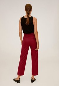 Vrouw met gevlochten haar in een zwart mouwloos top, rode high-waisted cropped broek en zwarte platte schoenen, staande tegen een effen achtergrond.