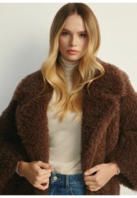 Brązowy, puszysty płaszcz o teksturowym wykończeniu, noszony na kremowym golfie. Modelka ma długie, blond włosy ułożone w luźne fale.