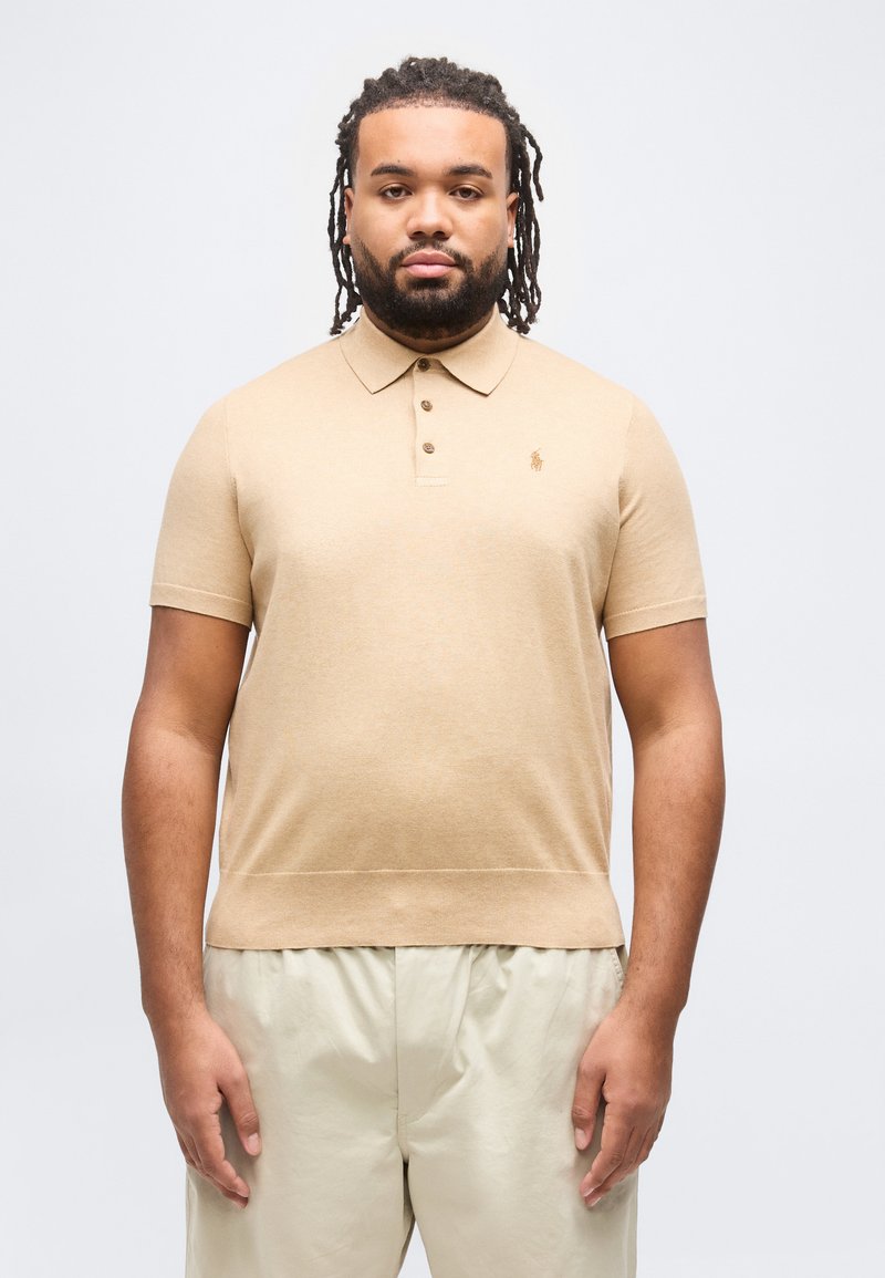 Άντρας με μεσαίου μήκους κόμπους μαλλιών, φοράει μπεζ κοντομάνικο πόλο πουκάμισο και ανοιχτόχρωμο παντελόνι, στέκεται μπροστά σε απλό φόντο.