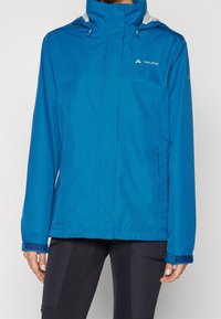 Veste imperméable bleue à coupe ajustée, avec capuche réglable et poches latérales zippées. Présente des accents contrastants gris et une texture de tissu.