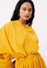 Chemise jaune à boutons, avec une coupe décontractée et une taille froncée. Manches longues et amples, en tissu lisse et léger.