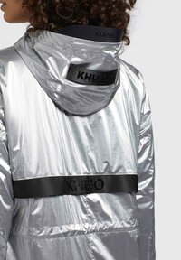 Strieborná metalická bunda s kapucňou, na zadnej strane s čiernym logom KHUJO a textúrovanou látkou, ktorá poskytuje hladký, reflexný vzhľad.
