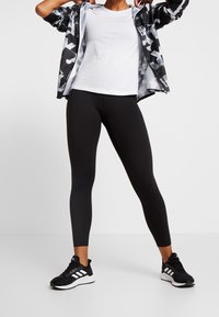 Leggings negras combinados con una chaqueta cortavientos gris de camuflaje y una camiseta blanca. La chaqueta cortavientos cuenta con un cierre de cremallera y telas contrastantes.