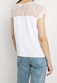Weißes Oberteil mit kurzen Spitzenärmeln und einer Schlüssellochöffnung hinten, aus strukturiertem Stoff und geradem Schnitt, kombiniert mit blauen Jeans.