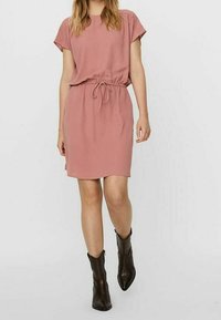 Růžové šaty s krátkým rukávem a staženým pasem, vyrobené z měkké látky a s volným střihem. Modelka nosí černé kotníkové boty pro kontrast.