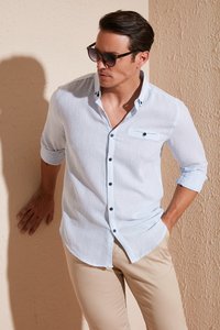 Lichtblauwe linnen blouse met lange mouwen, zwarte knopen en een borstzak, gecombineerd met beige broek. Gladde textuur tegen een gestructureerde muur.