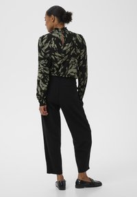 Zwarte broek met hoge taille, gecombineerd met een blouse met donkergroene abstracte bladprint. De blouse heeft een gerufflede kraag en een knoopdetail aan de achterkant.