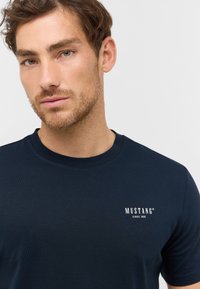 T-shirt bleu marine à col rond en coton. Présente un logo blanc "MUSTANG" avec le texte "SINCE 1932" sur la partie gauche de la poitrine. Texture lisse.