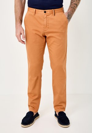 Man wearing slim-fit orange pants, navy loafers, and a navy shirt, standing with hands in pockets against a plain background.