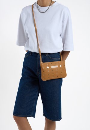 Tan synthetische schoudertas met studs en gaasdetails, rechthoekige vorm, verstelbare schouderband en een gladde textuur. Draagt bij casual denim shorts.